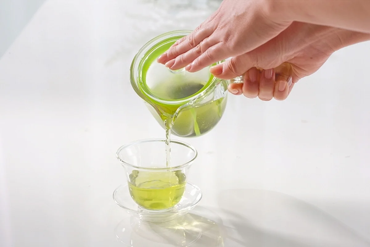 ハリオ 耐熱ガラス茶器 ─ 透明な急須からガラス杯へ淹れたての緑茶を注ぐ手元、葉茶が湯に開く