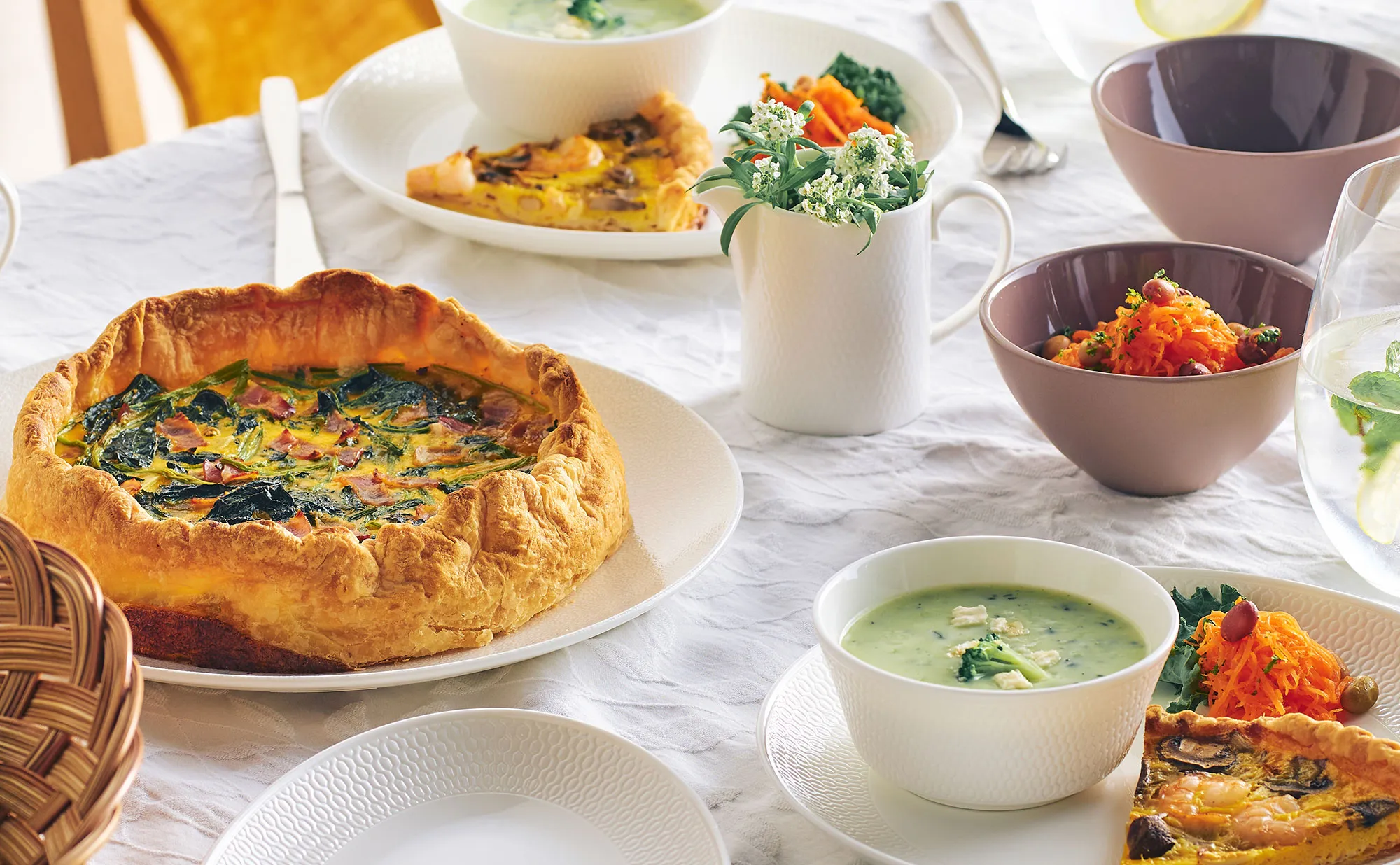 白い食器を並べた食卓：キッシュやスープ、キャロットラペ、花を活けた白い花瓶、ピンクのカップと籠