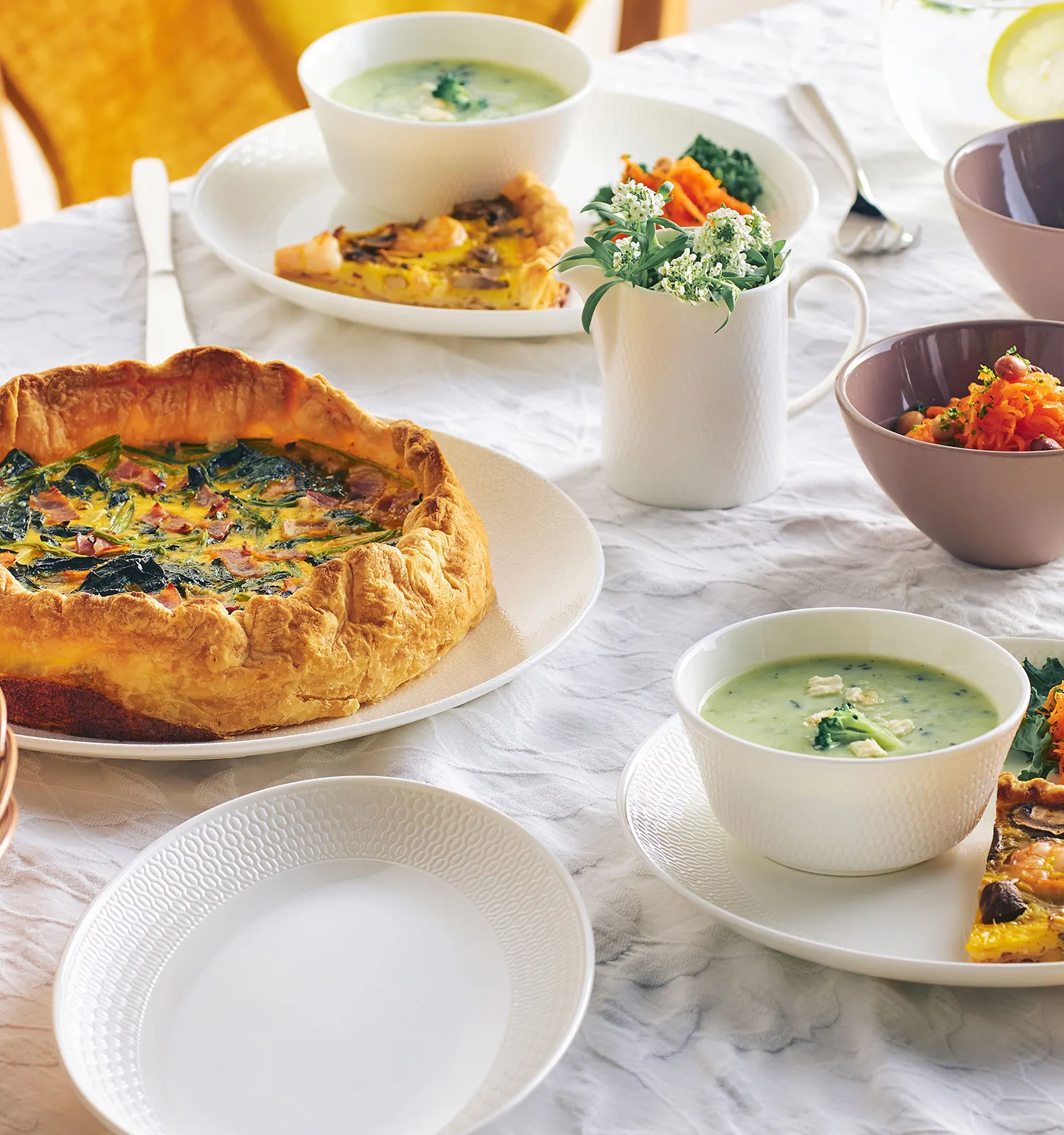 白い食器を並べた食卓：キッシュやスープ、キャロットラペ、花を活けた白い花瓶、ピンクのカップと籠